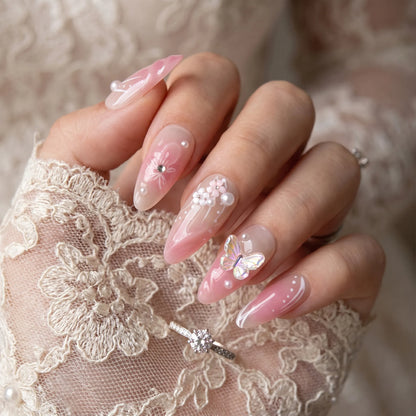 Garden of Forever pink sheer nail art with holographic 3D butterfly, white floral clusters, pearl embellishments and swirl details on almond nails, styled with diamond ring on lace