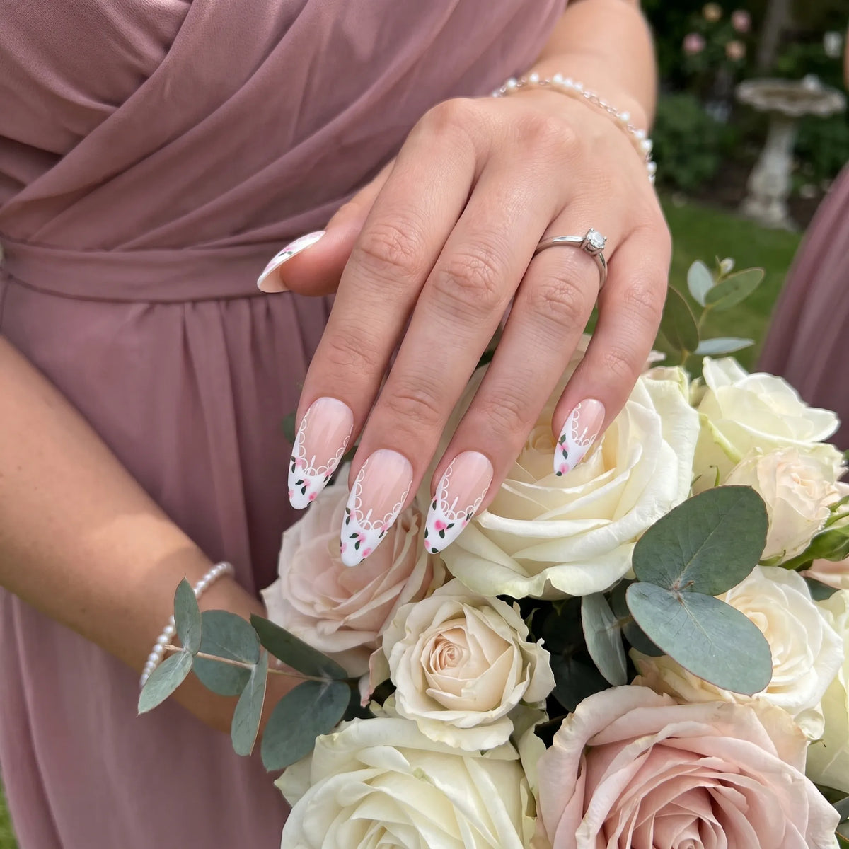 Ivory Bloom French bridal nail art with white lace scallop French tips and pink rose floral accents on almond nails, worn with diamond engagement ring against blush rose bouquet