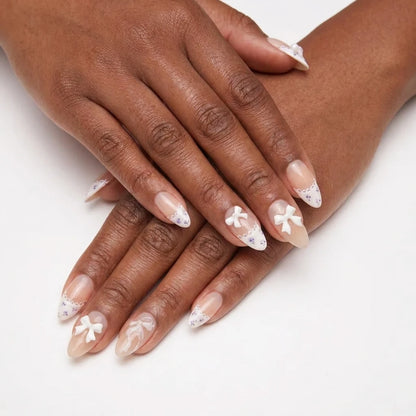 Ribbon Muse sheer nude nail art with 3D white bow charms, lace scalloped French tips and tiny lavender floral patterns on almond nails against white background