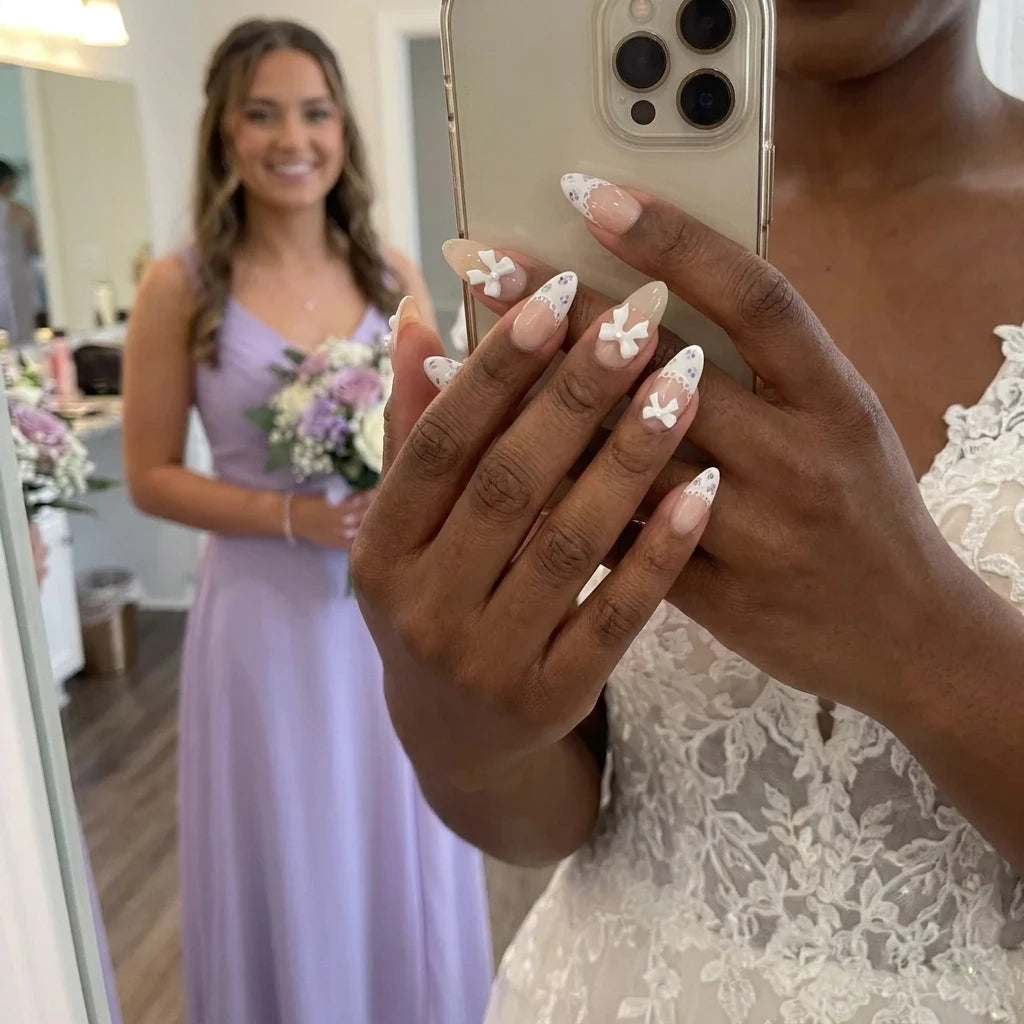 Ribbon Muse bridal nail art with 3D white bow charms, lace scallop French tips and tiny purple floral accents on almond nails, mirror selfie with bridesmaid in lavender dress