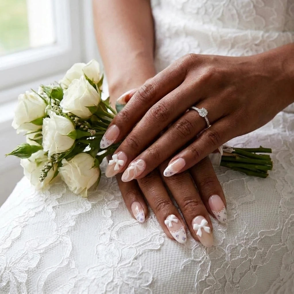 Ribbon Muse bridal nail art with 3D white bow, scalloped lace French tips and purple floral details on almond nails, styled with white rose bouquet and diamond engagement ring